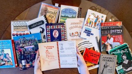 Hands holding open a vintage cookbook with a number of other cookbooks in the background.