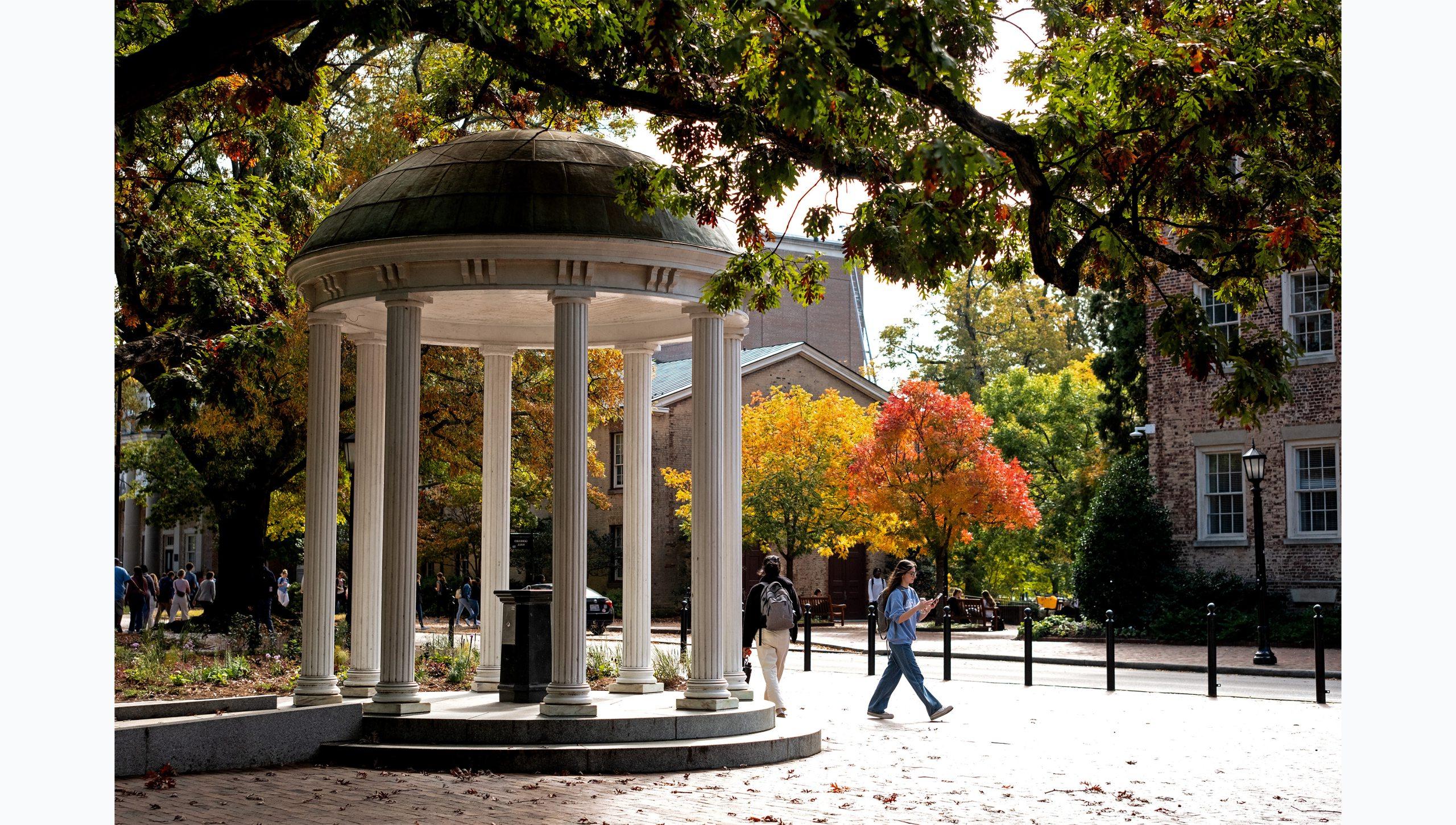 A student walks by the Old Well on the campus of UNC-Chapel Hill. Trees with red, yellow and green leaves are seen in the background.